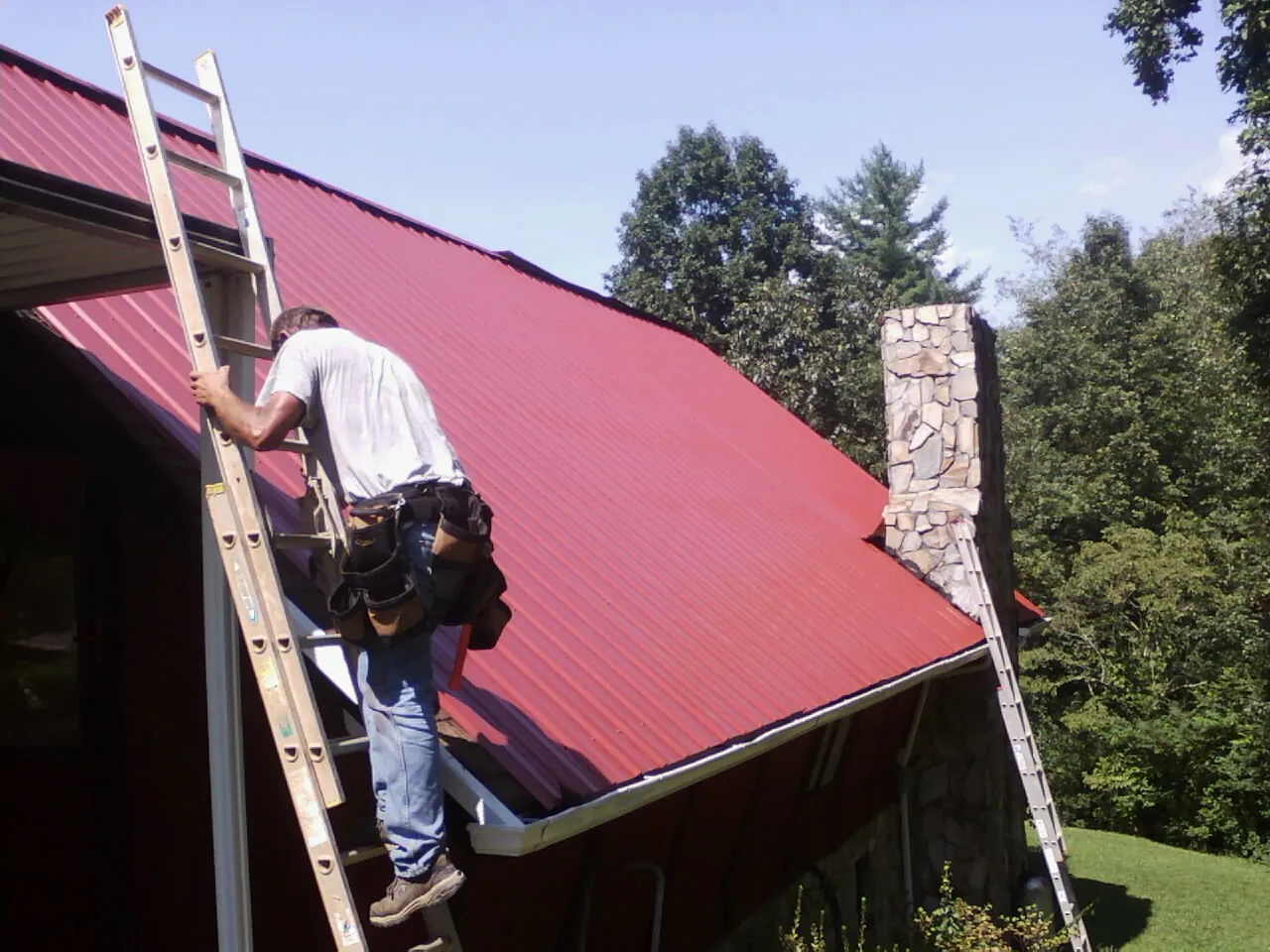 Roofer on a ladder beside a red metal roof and stone chimney — Soffit & Fascia Repair by Roofing Brothers in New Oxford